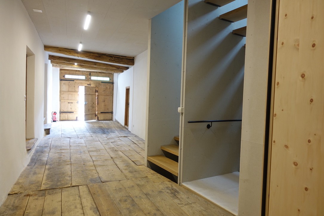 Blick in den historischen Suler mit dem neuen offen Treppenraum. Ein neues Oberlicht erhellt die offene zweiläufige Holztreppe vom Obergeschoss bis in den Keller. Die neuen Werkstoffplatten übernehmen die Rohheit der gekalkten Bruchsteinwände und des alten Holzbodens. 