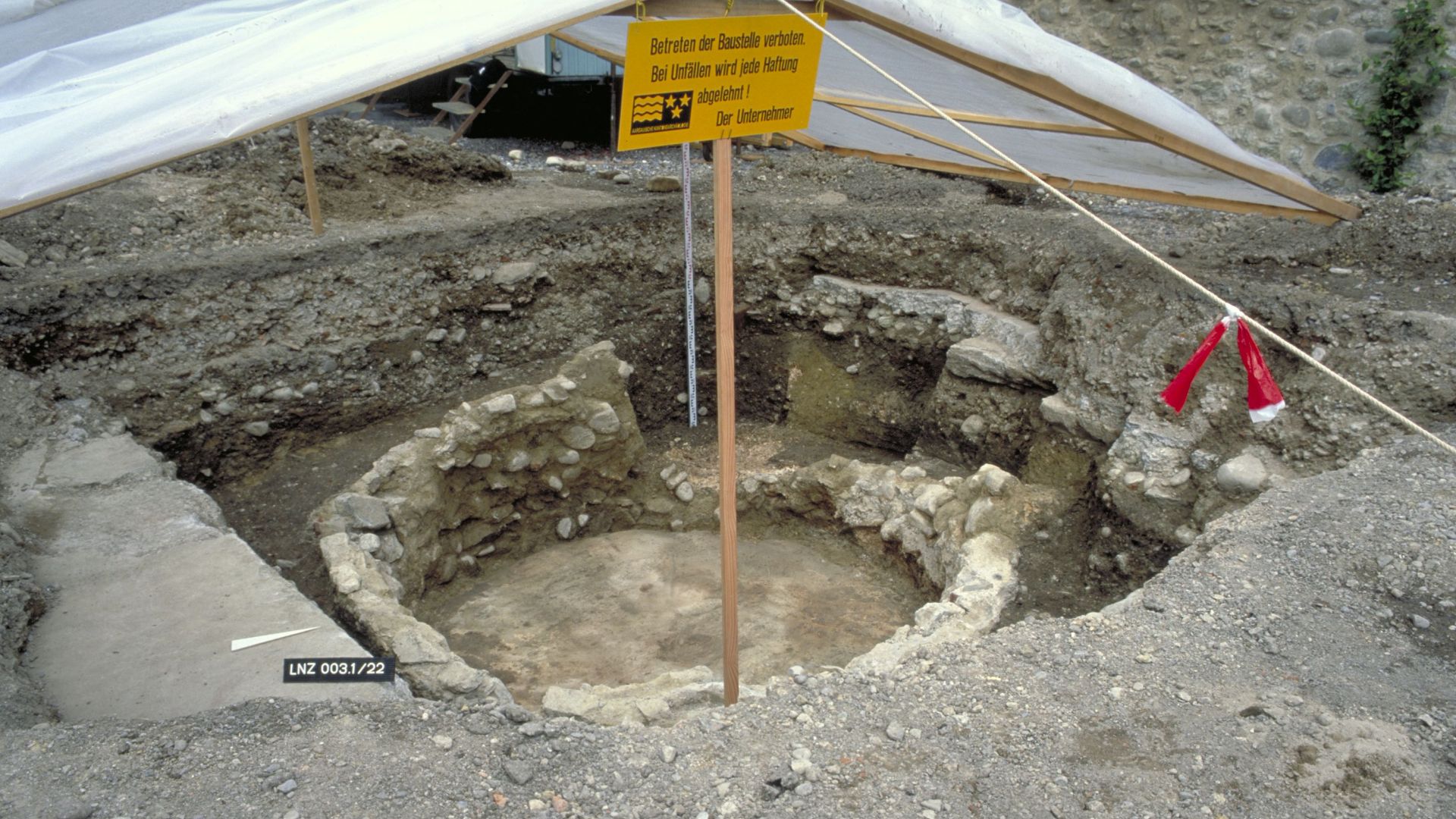 Runde Struktur aus Steinen direkt unter der Oberfläche, mit Baustellenschild versehen.  